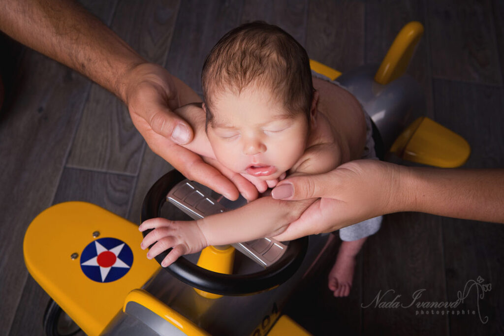 Un nouveau-né endormi repose dans les bras de ses parents, délicatement bercé par un avion jouet jaune. Ses yeux sont fermés et sa peau est douce. La scène est réconfortante et apaisante.