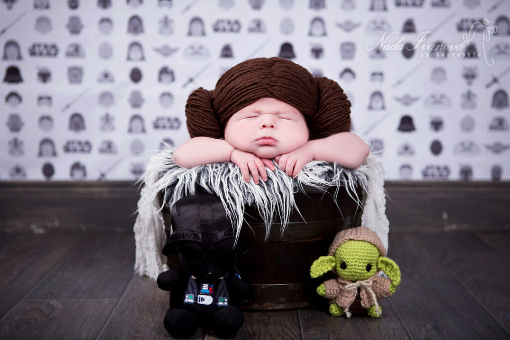 Un adorable nouveau-né dort profondément, coiffé d'un chapeau marron inspiré de la Princesse Leia, blotti dans un seau en bois rustique doublé d'une douce fourrure blanche. Un amigurumi de Dark Vador et Yoda est posé à ses côtés, complétant ainsi la séance photo de nouveau-né sur le thème de Star Wars.