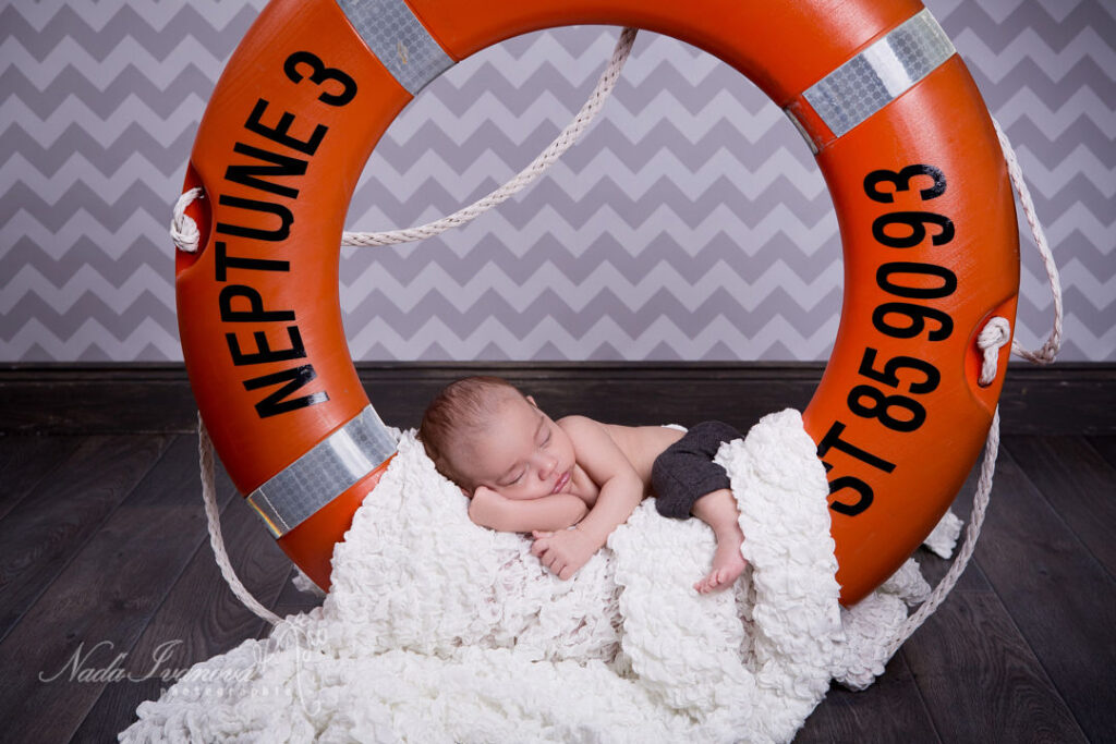 Un nouveau-né paisible dort profondément niché dans une couverture blanche au crochet, partiellement caché derrière un grand gilet de sauvetage orange marqué « NEPTUNE 3 » et « 07 85 90 93 ».