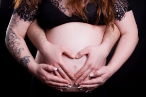 Gros plan sur le ventre d'une femme enceinte, orné d'un cœur formé par ses mains et celles de son partenaire. La femme porte un haut en dentelle noire et son bras arbore un tatouage floral. L'image évoque l'amour et l'attente de la grossesse.