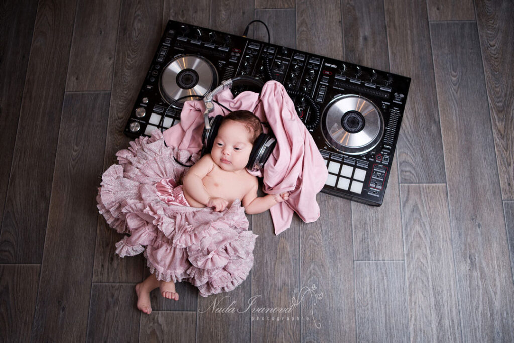 Une adorable petite fille en tutu rose, portant un casque audio surdimensionné, est allongée sur le parquet à côté d'une table de mixage. Elle regarde paisiblement la caméra.