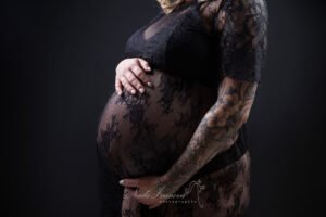 Comment s’habiller quand on est enceinte ? Gros plan sur le ventre d'une femme enceinte, délicatement bercé par ses mains. Elle porte une robe de maternité en dentelle noire transparente, son bras orné de tatouages délicats. Le fond sombre souligne la douce luminosité de sa peau.