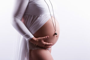 Photographe grossesse Sète Gros plan sur le ventre d'une femme enceinte, orné d'un bola de grossesse, un délicat pendentif en argent sur un cordon noir. Elle enveloppe délicatement son ventre rond, vêtue d'une robe de maternité blanche fluide et de sous-vêtements en dentelle. L'arrière-plan est d'un blanc doux et lumineux.
