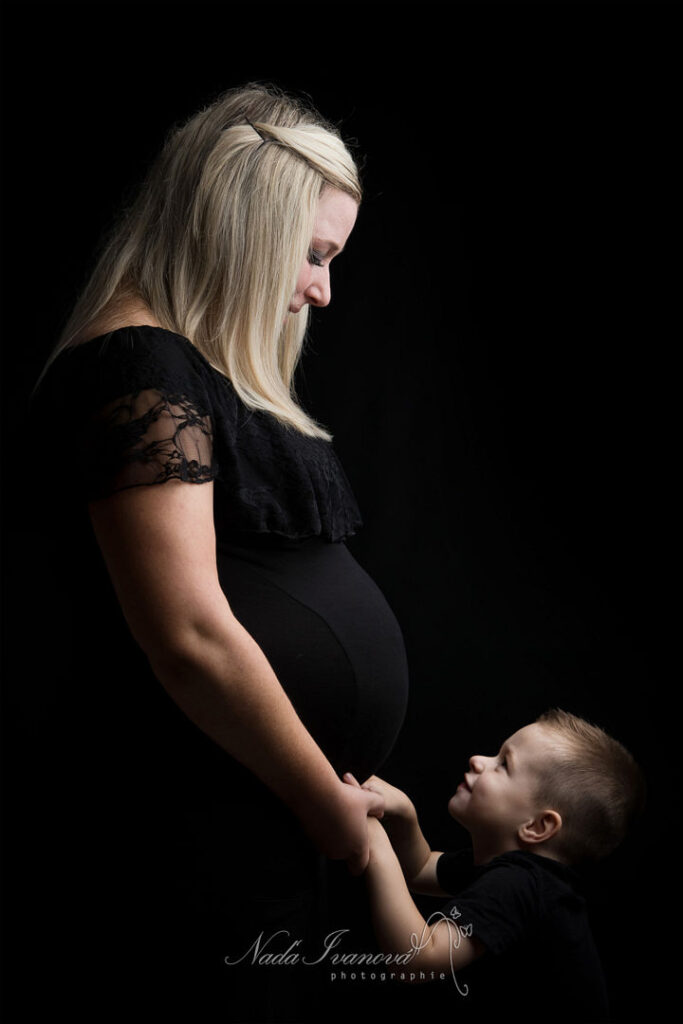Une femme blonde enceinte, vêtue de noir, regarde tendrement son fils. L'enfant, également en noir, lui tient la main, les yeux levés vers elle. Photo de grossesse capturant un moment précieux de famille.