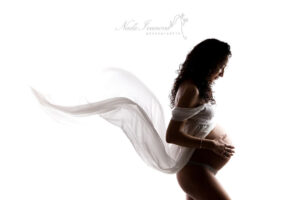 Silhouette d'une femme enceinte vêtue d'un tissu blanc fluide, les mains caressant délicatement son ventre rond. L'image évoque un sentiment de sérénité et d'impatience.