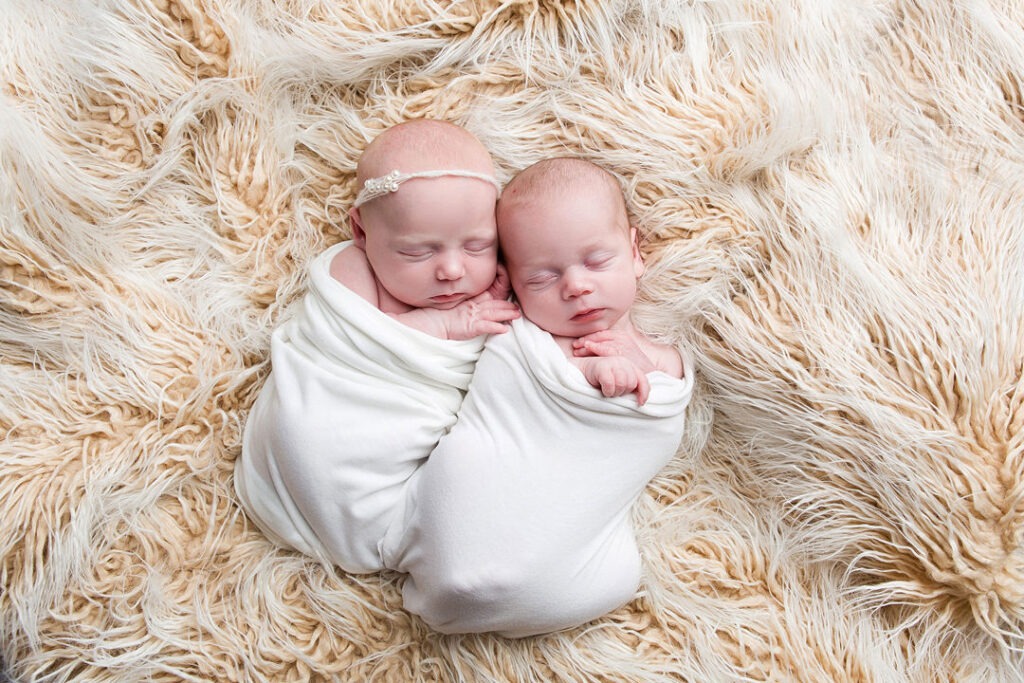 Deux adorables bébés jumeaux emmaillotés dans des couvertures blanches, dormant paisiblement sur une douce couverture duveteuse beige. L'un porte un délicat bandeau à perles.