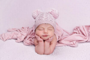 Un nouveau-né dort le menton posé sur les mains, les coudes pliés et le visage tourné vers l'avant. Le bébé porte un bonnet en tricot rose pâle avec deux pompons et est enveloppé dans une couverture rose douce sur une surface texturée assortie.