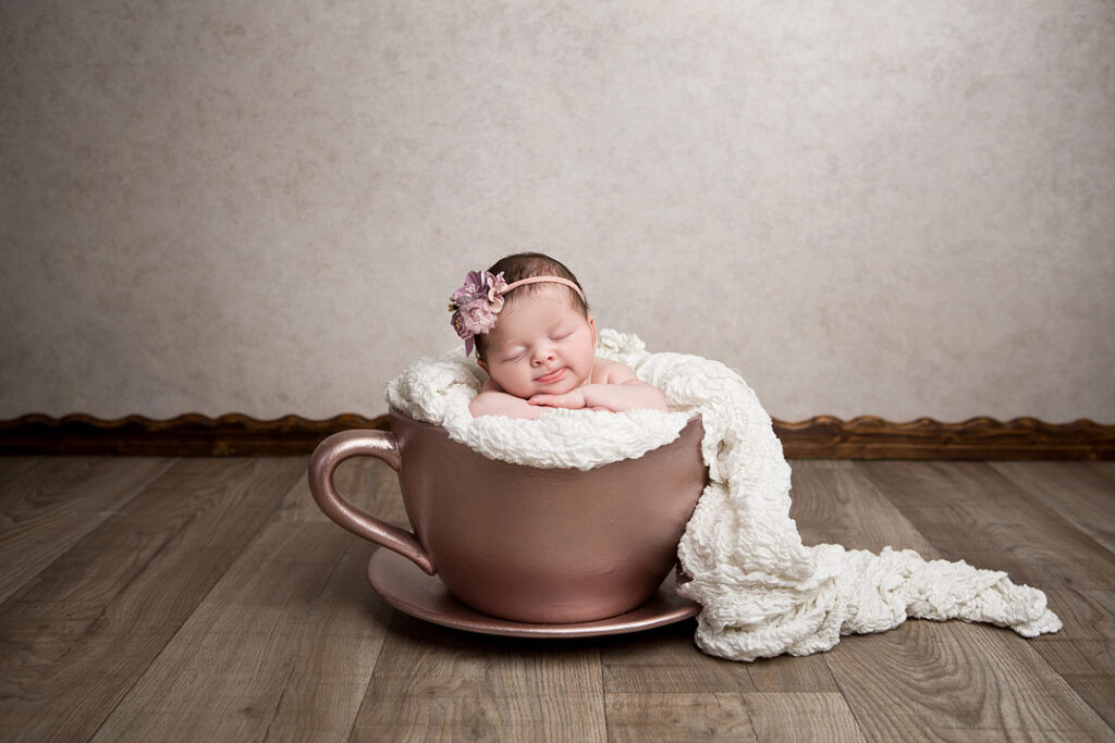 Une petite fille nouveau-née, coiffée d'un délicat bandeau fleuri, dort profondément blottie dans une grande tasse à thé rose doré. Elle est emmaillotée dans une douce couverture blanche au crochet qui s'étale sur le parquet. La scène paisible se détache sur un mur aux tons neutres.