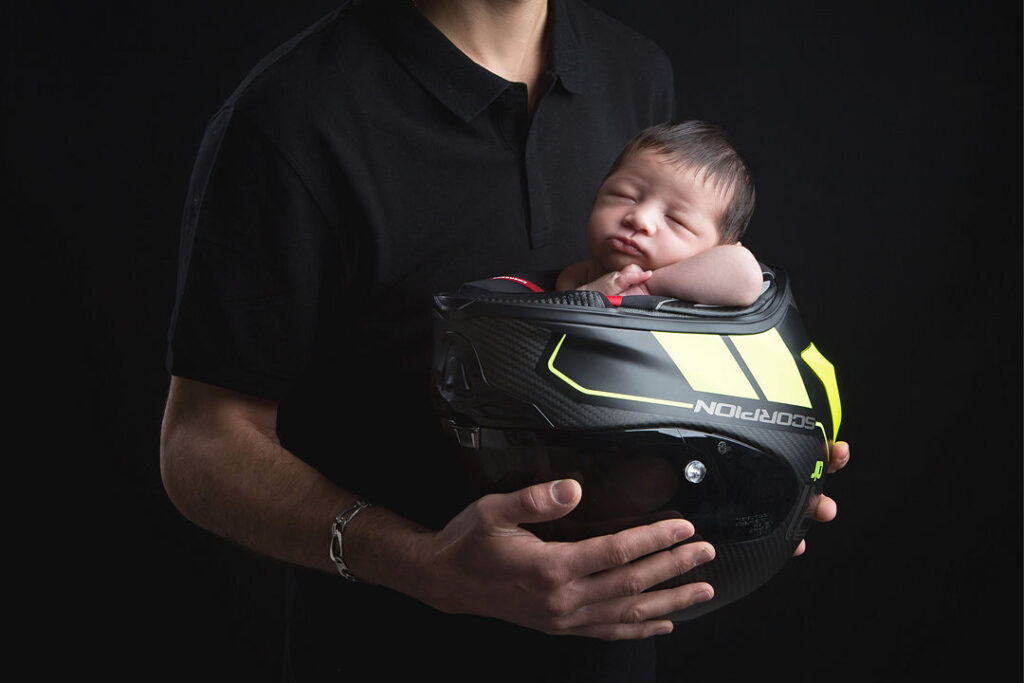 Un nouveau-né dort profondément, blotti dans un casque de moto Scorpion noir et jaune fluo, bercé par les mains d'un homme vêtu d'un polo noir. Le fond sombre souligne le contraste entre le casque et les traits délicats du bébé.