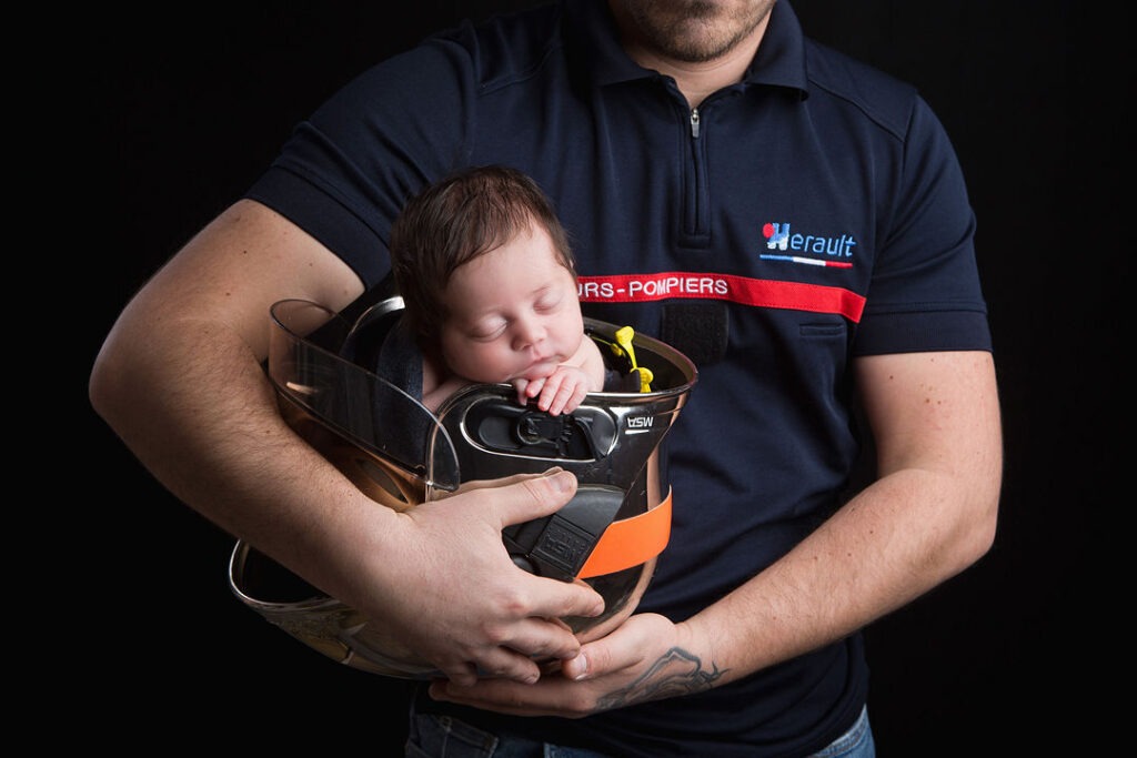 Un nouveau-né dort paisiblement blotti dans un casque de pompier, bercé dans les bras musclés d'un pompier vêtu d'un uniforme bleu foncé « Herault - Pompiers ». Cette scène touchante se détache sur un fond noir.