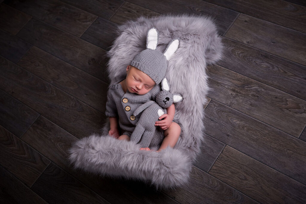 Un nouveau-né, endormi, blottit contre un lapin tricoté, vêtu d'une adorable tenue et d'un bonnet gris. Il repose sur un tapis gris moelleux, posé sur un parquet sombre.
