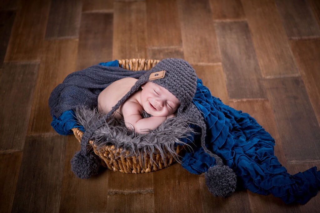 Un nouveau-né souriant dort profondément dans un panier en osier, blotti dans un bonnet gris et une couverture moelleuse. Il est entouré d'un tissu à volants bleu sarcelle sur un parquet. Une jolie photo de nouveau-né.