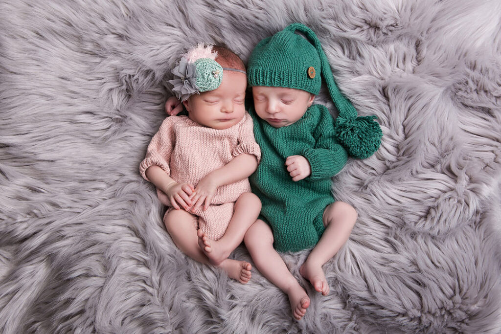 Deux jumeaux nouveau-nés sont blottis l'un contre l'autre sur une couverture grise et moelleuse. L'un porte une tenue en tricot rose et un bandeau fleuri, tandis que l'autre porte une barboteuse verte et un bonnet assorti. Ils dorment paisiblement.