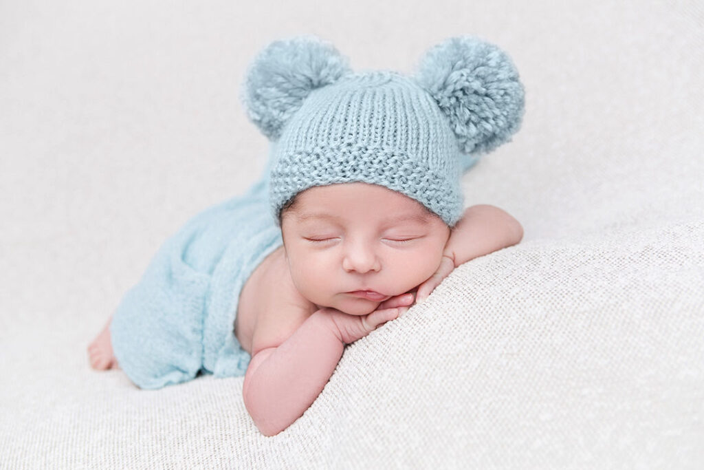 Un adorable petit garçon nouveau-né dort paisiblement sur une douce couverture blanche, coiffé d'un bonnet bleu clair en tricot orné de pompons moelleux. Ses mains sont délicatement glissées sous son menton.