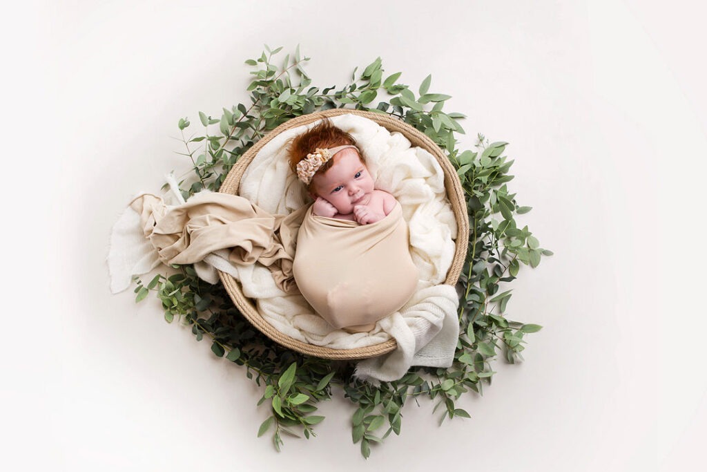 Un nouveau-né, une petite fille aux cheveux roux éclatants, coiffé d'un bandeau fleuri, est enveloppé dans un tissu beige à l'intérieur d'un panier tressé rustique. Une luxuriante guirlande de feuilles d'eucalyptus entoure le panier, créant une scène photographique de nouveau-né sereine et naturelle.