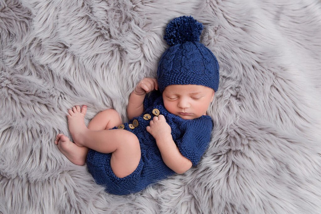 Un nouveau-né paisible dort profondément sur un tapis gris moelleux, vêtu d'une adorable barboteuse bleue tricotée main et d'un bonnet torsadé assorti à pompon. Son expression douce est visible, et l'ambiance générale est sereine et réconfortante.