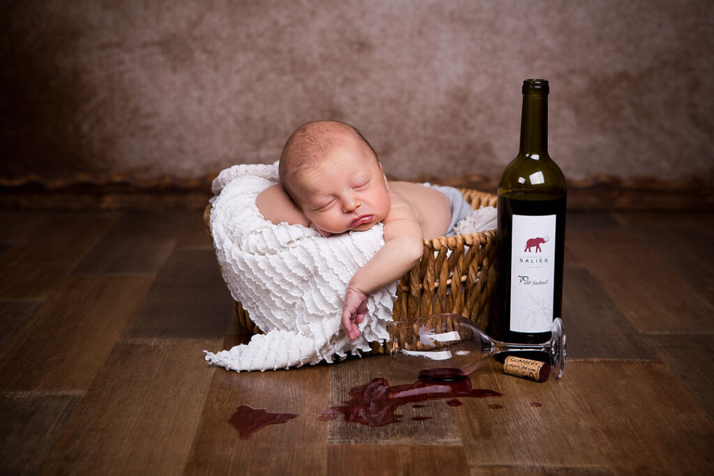 Un nouveau-né dort profondément dans un panier en osier doublé de tissu blanc, à côté d'un verre de vin rouge renversé et d'une bouteille de Petit Pastisoul de Salies. La scène se déroule sur un parquet sombre, devant un mur marron texturé.