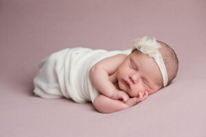 Réussir la séance photo de votre bébé de 2 mois en studio Un nouveau-né dort paisiblement, emmailloté dans un châle couleur crème, la tête posée sur son bras, sur une douce couverture rose. Il porte un délicat bandeau à fleurs blanc cassé. L'image évoque la sérénité et une nouvelle vie.
