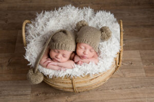 Réussir votre séance photo jumeaux : conseils et astuces Deux jumeaux nouveau-nés se blottissent l'un contre l'autre, dormant profondément dans un panier en osier doublé d'une douce fourrure blanche. Ils portent d'adorables bonnets en tricot assortis à pompons, créant une ambiance apaisante et réconfortante.