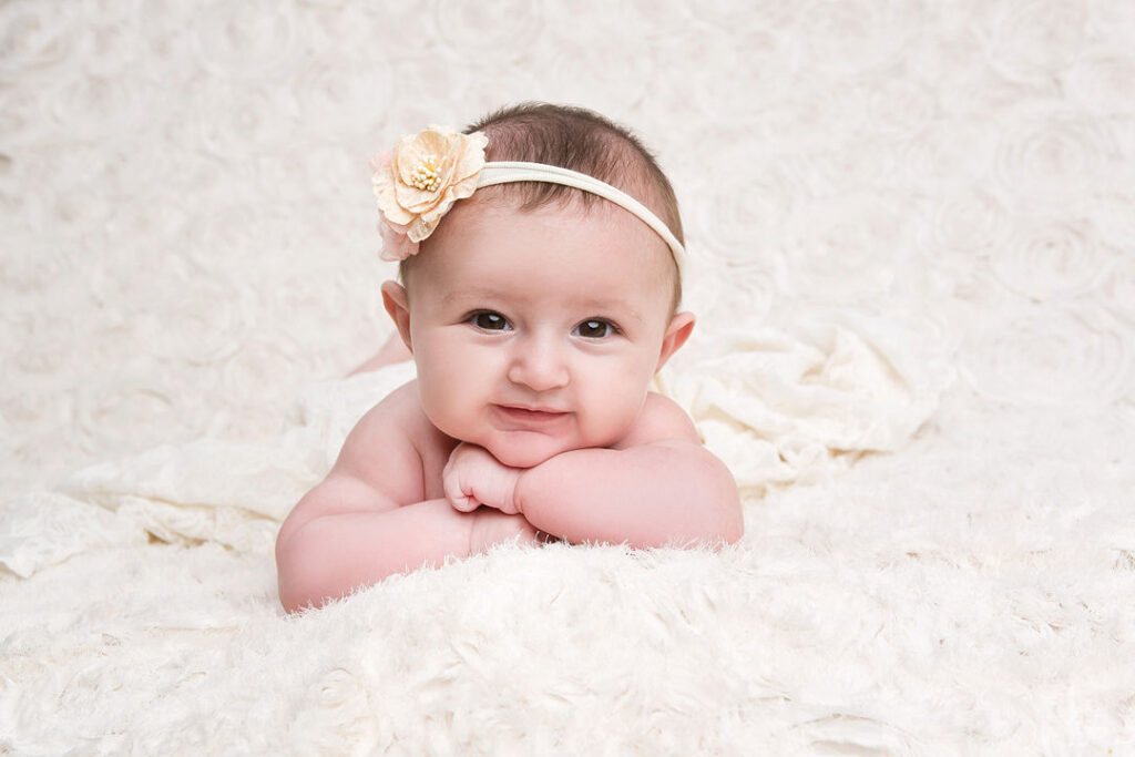 Adorable petite fille aux cheveux noirs, portant un bandeau fleuri beige, allongée sur le ventre sur une couverture blanche moelleuse, souriant doucement à la caméra.