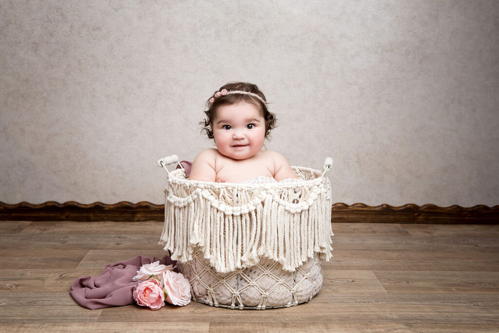 Une adorable petite fille ronde aux cheveux bruns bouclés est assise dans un panier en macramé couleur crème, coiffée d'un serre-tête à fleurs roses. Elle regarde l'objectif droit dans les yeux avec une expression douce. Le panier est posé sur un parquet contre un mur aux tons neutres. Des roses roses et un tissu mauve rehaussent la scène.