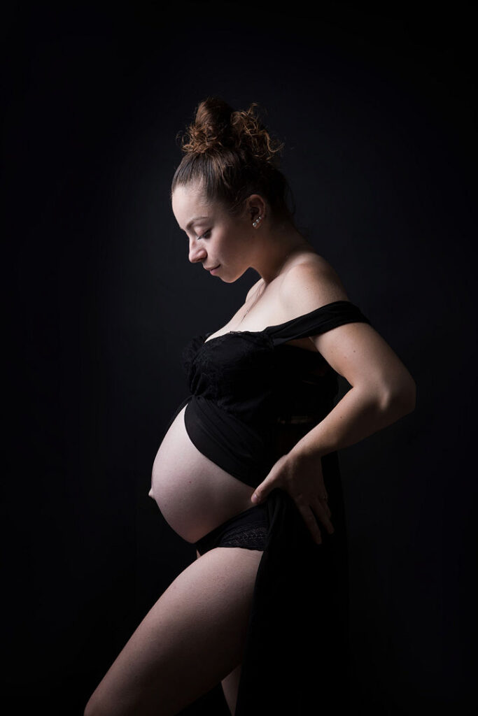 Une femme enceinte, en robe noire, pose de profil dans un studio sombre. Elle regarde son ventre arrondi, capturant la beauté de la grossesse. Photo de grossesse.