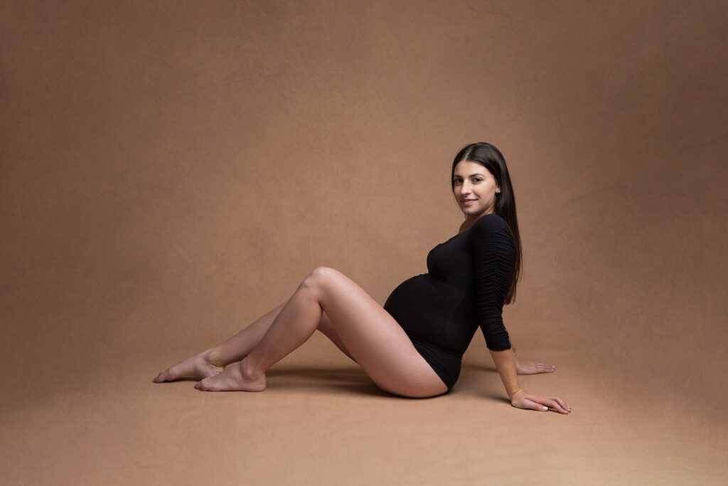 Une femme enceinte, vêtue d'un body noir, pose sereinement sur un fond marron uni. Elle sourit, mettant en valeur son ventre arrondi, pour une séance photo de maternité.