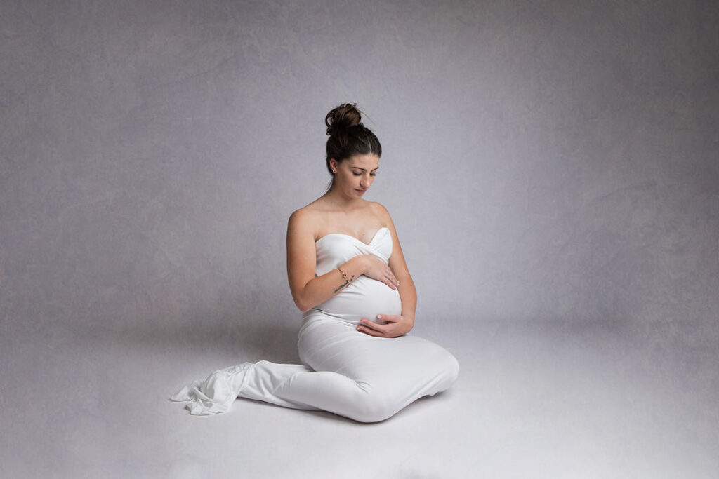 Future maman rayonnante dans une robe blanche, posant délicatement. Séance photo grossesse en studio, tenant son ventre arrondi. La lumière douce met en valeur la beauté de la maternité.