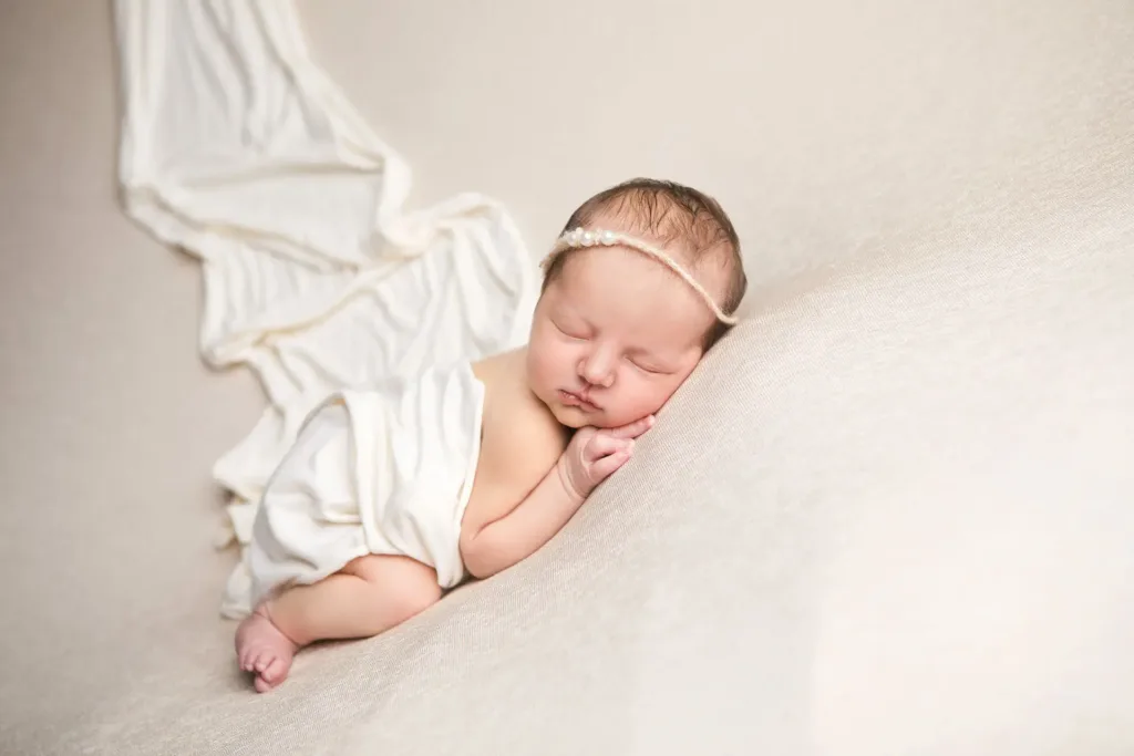 Un nouveau-né, une petite fille, dort paisiblement, emmailloté dans un tissu crème, sur une surface texturée beige. Il porte un délicat bandeau de perles et ses mains sont délicatement glissées sous son menton. Un léger châle crème flotte derrière elle.