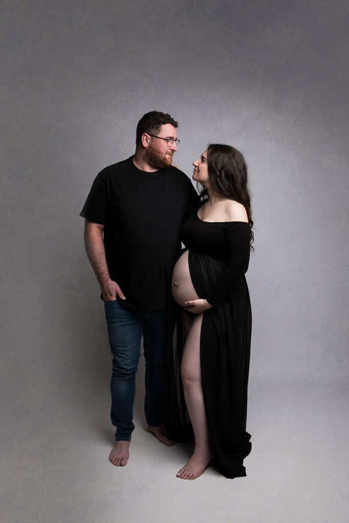 Un couple enceinte, la future maman portant une robe noire élégante, et le futur papa en t-shirt noir et jean, se regardent avec amour. Séance photo de grossesse en studio, ambiance douce.