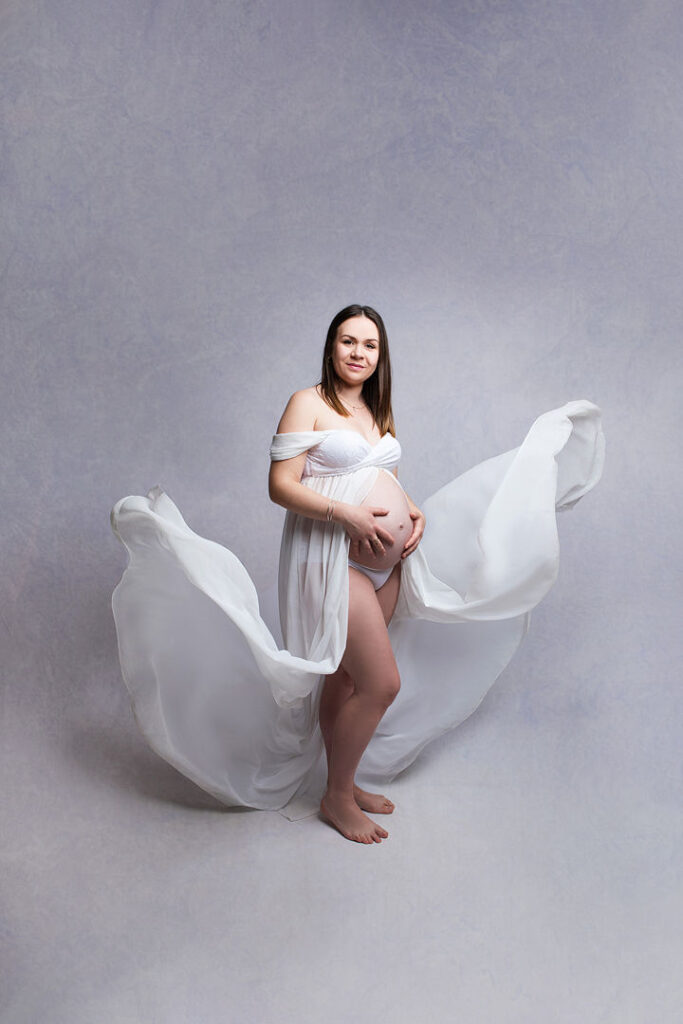 Future maman rayonnante en robe blanche, tenant son ventre arrondi, posant pour une photo de grossesse. Une étoffe vaporeuse flotte autour d'elle, créant un effet de mouvement et de douceur. Séance photo de maternité.