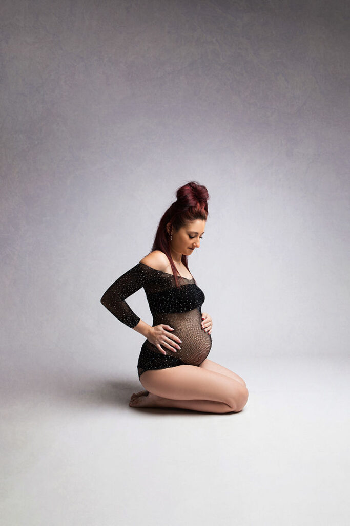 Future maman agenouillée, portant une combinaison noire transparente ornée de paillettes, caressant son ventre rond. Cheveux roux attachés, posant devant un fond gris clair pour une séance photo de maternité.