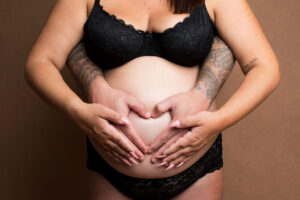 Gros plan d'une femme enceinte et des mains de son partenaire formant un cœur sur son ventre rond. Elles portent toutes deux de la lingerie en dentelle noire sur un fond marron. Le bras de la femme est tatoué, celui de l'homme, un autre sur la manche. L'image évoque l'amour, la famille et l'attente de la parentalité.