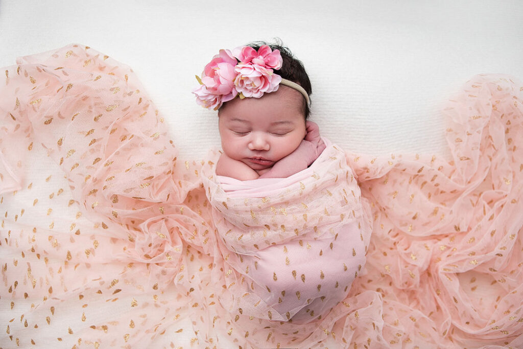 Nouveau-né dormant paisiblement, enveloppé dans un tissu rose délicat orné de motifs dorés, coiffé d'un bandeau floral rose. Douceur et innocence capturées.