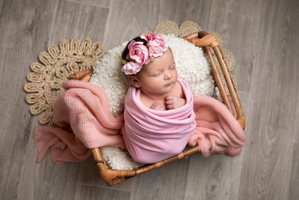 Une petite fille nouveau-née dort profondément dans un panier en osier, blottie dans un lange rose et ornée d'une délicate couronne de fleurs roses. Elle repose sur un coussin blanc moelleux, enveloppée d'une couverture couleur pêche. La scène est paisible et sereine.