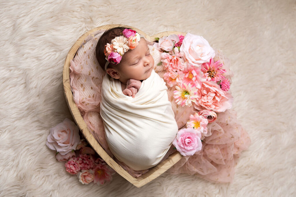 Un nouveau-né, enveloppé dans un tissu crème et coiffé d'une couronne de fleurs roses, dort paisiblement dans un panier en bois en forme de cœur. Autour d'elle, de douces fleurs roses et d'un tulle rose poudré créent une scène photographique de nouveau-né douce et onirique.