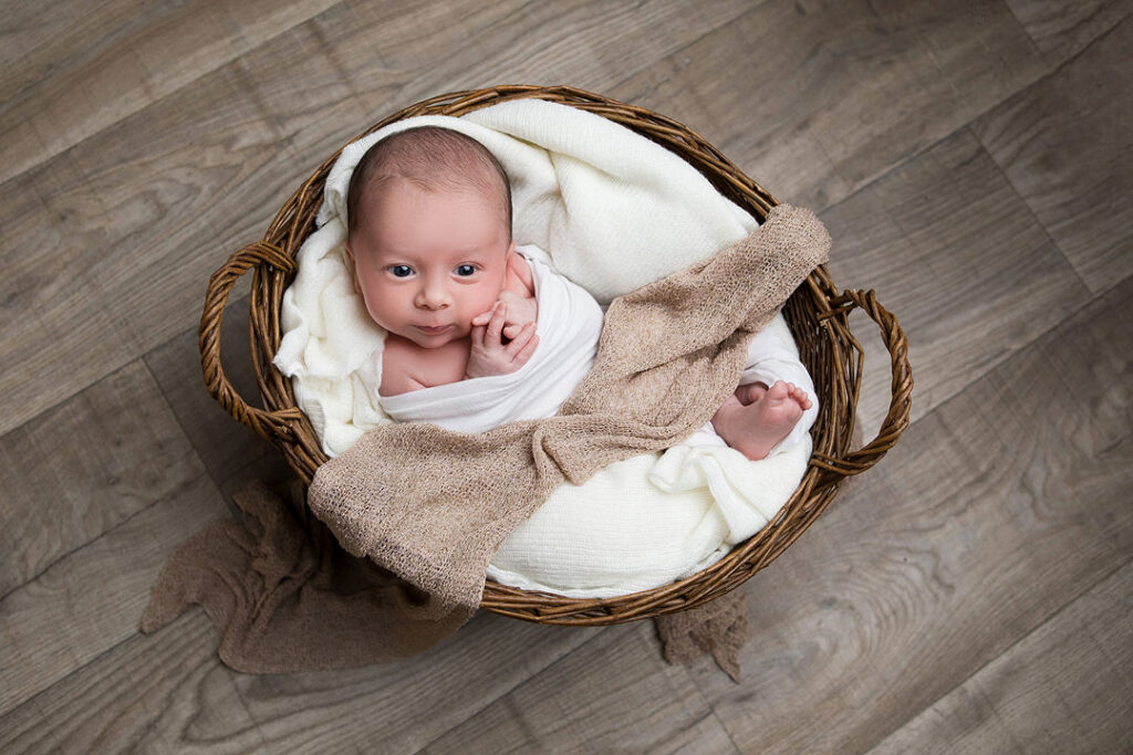Adorable nouveau-né blotti dans un panier en osier rustique, emmailloté dans de doux tissus blancs et beiges. Son expression paisible et ses joues rebondies sont captivantes. Une charmante photo de nouveau-né.