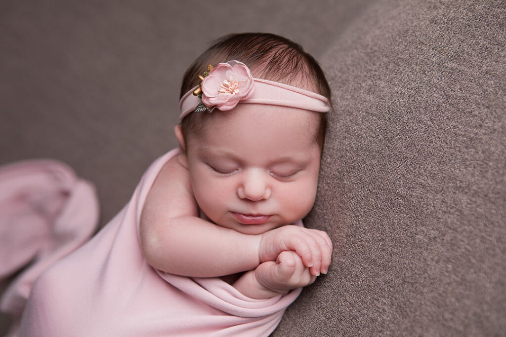 Un nouveau-né dort paisiblement, emmailloté dans un doux châle rose, la tête appuyée contre un tissu taupe. Il porte un délicat bandeau rose orné d'une petite fleur. L'image évoque la douceur et l'innocence du nouveau-né.