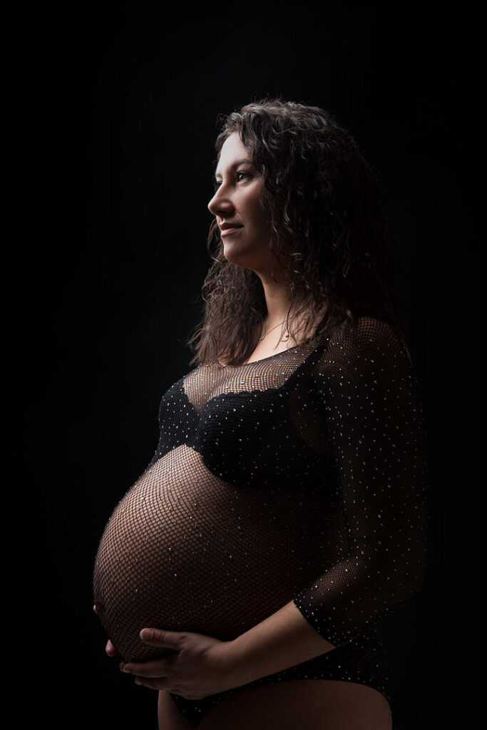 Une femme enceinte radieuse aux cheveux noirs et bouclés regarde pensivement à ses côtés, berçant son ventre rond. Elle porte un body noir en maille scintillante sur un fond sombre, créant un portrait de maternité à la fois dramatique et intime.