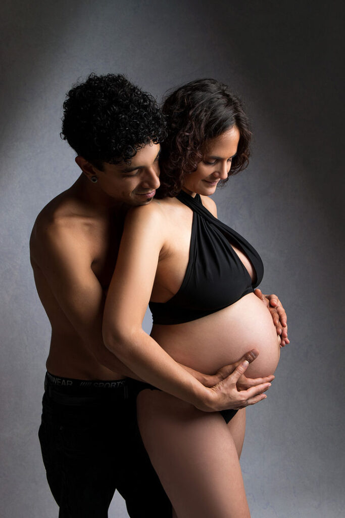 Un couple amoureux s'enlace lors d'une séance photo de maternité, le ventre de la femme enceinte étant bien visible. L'homme, torse nu, berce son ventre, tous deux souriant d'impatience. Une lumière tamisée et un fond gris créent une ambiance tendre, parfaite pour célébrer les futurs parents.
