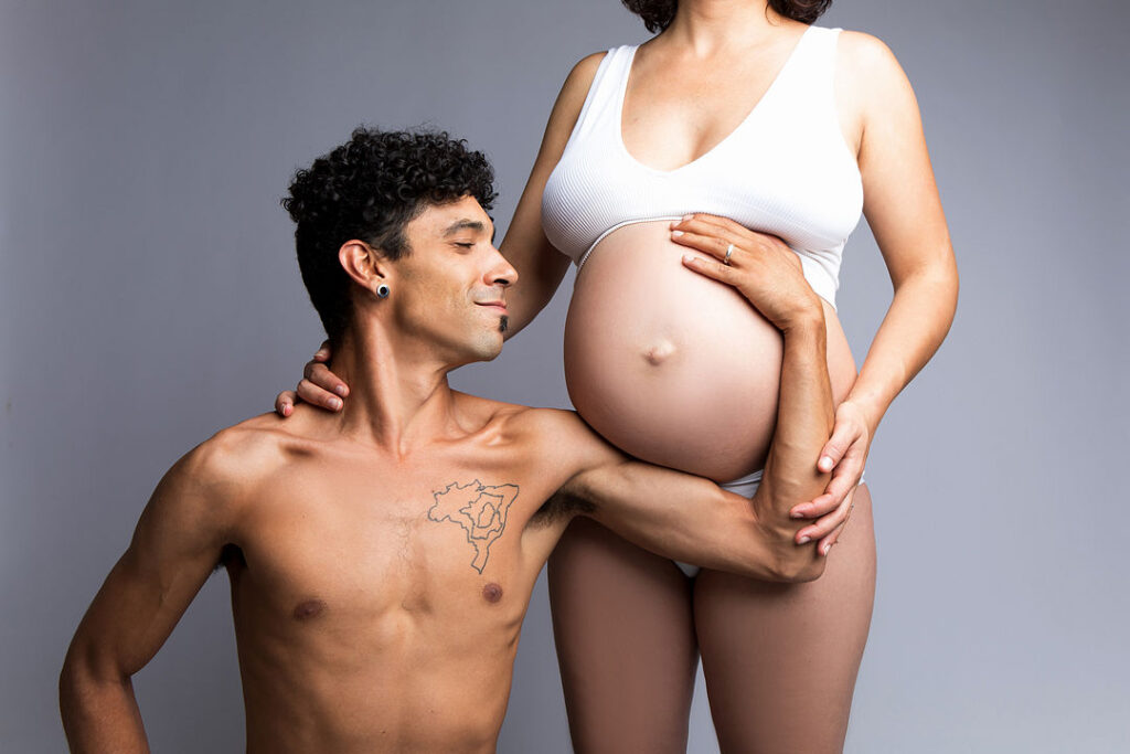 Un homme torse nu aux cheveux bouclés contemple avec adoration sa compagne enceinte, vêtue d'un débardeur blanc et tenant son ventre. Leurs mains sont entrelacées, célébrant la joie de la grossesse et de l'arrivée du bébé.