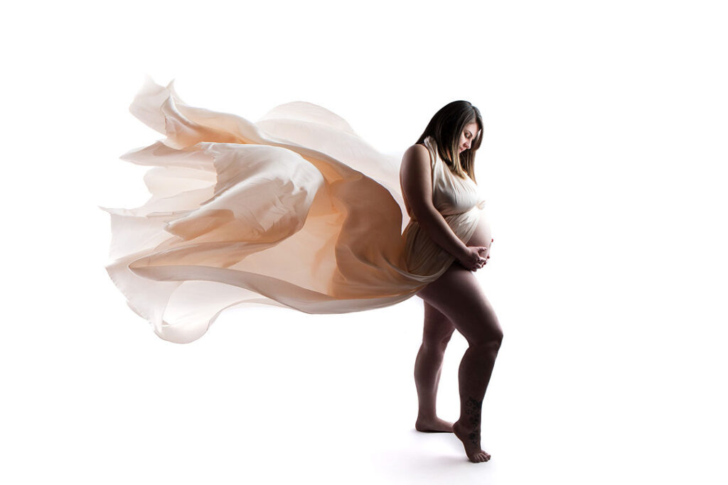 Une femme enceinte, vêtue d'une robe fluide couleur crème, enveloppe son ventre lors d'une magnifique séance photo de maternité. Le tissu ondule autour d'elle, créant un effet spectaculaire pour les photos de maternité.