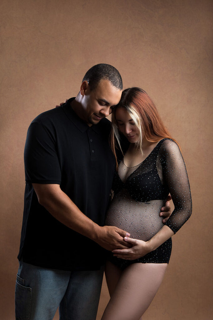 De futurs parents s'enlacent lors d'une séance photo de maternité, le ventre rond de la mère se dessinant sous un body noir transparent. Le père, vêtu d'un polo noir, berce doucement son ventre tandis qu'ils se contemplent avec amour, sur fond brun chaleureux, incarnant la joie de la grossesse.