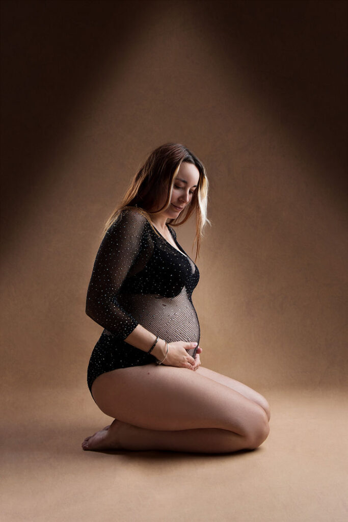 Une femme enceinte est agenouillée sur un fond marron, berçant son ventre rond. Elle porte un body noir transparent orné de paillettes, ses cheveux tombant en cascade sur ses épaules. Une belle idée de séance photo de maternité.