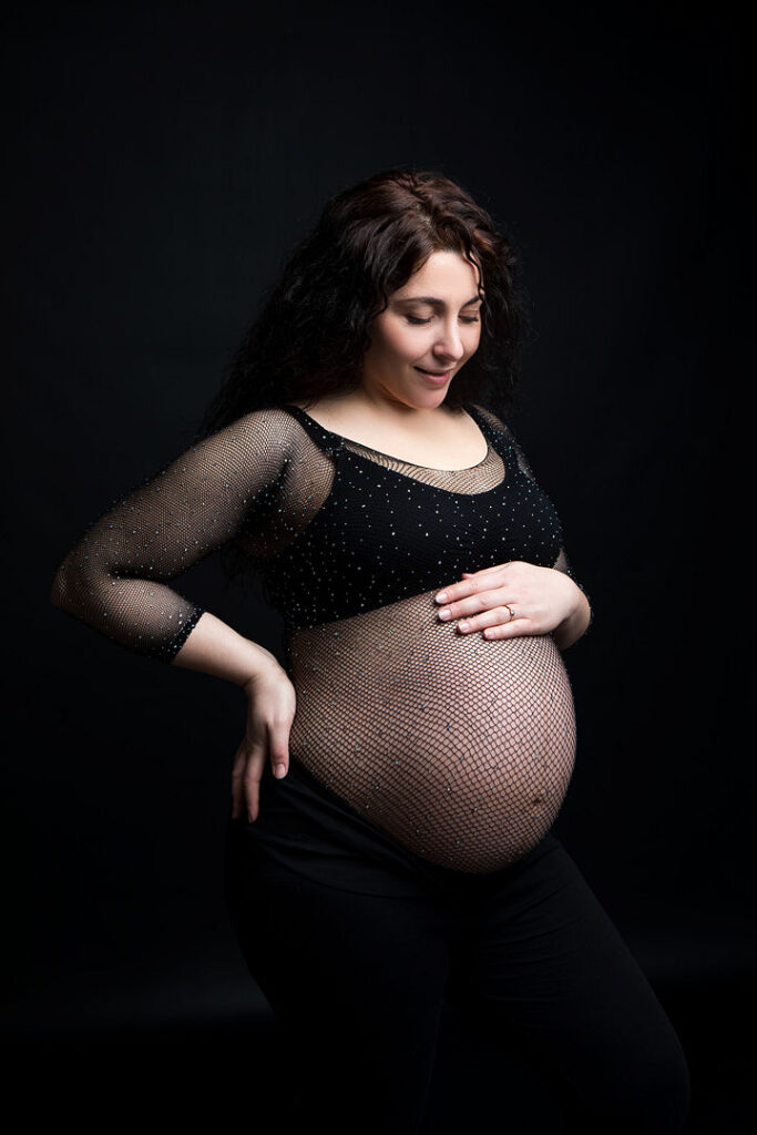 Une belle femme enceinte, vêtue d'un body noir et d'un haut résille, berce tendrement son ventre rond. Une photographie de maternité saisissante sur fond noir souligne la joie de la future maman.