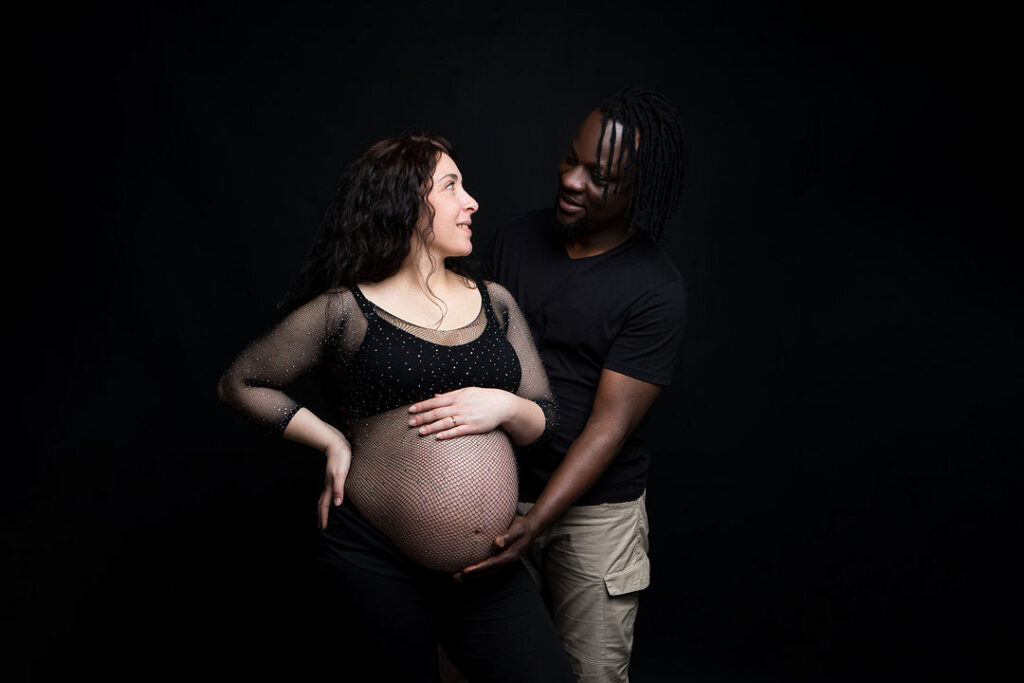 De futurs parents échangent un regard tendre lors d'une séance photo de maternité. La future maman, vêtue d'une tenue noire et d'un haut en résille, berce son ventre tandis que son partenaire le caresse délicatement. Fond noir.