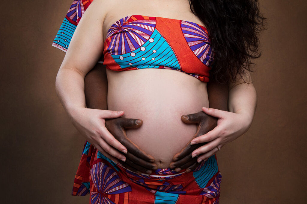 Une femme enceinte, vêtue d'un haut et d'une jupe à imprimé africain éclatant, est enlacée par son partenaire, dont les mains forment un cœur sur son ventre. Ce couple, symbole de mixité, est mis en valeur dans cette tendre séance photo de maternité, symbole d'amour et de famille.