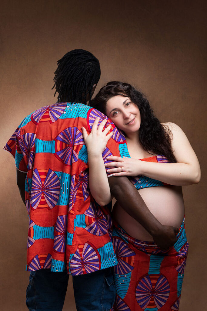 Une femme enceinte sourit, appuyée contre son partenaire, enlaçant son ventre rond. Ils portent des tissus africains assortis pour une séance photo de maternité, célébrant leur statut de couple interracial et l'espoir d'une famille diversifiée.
