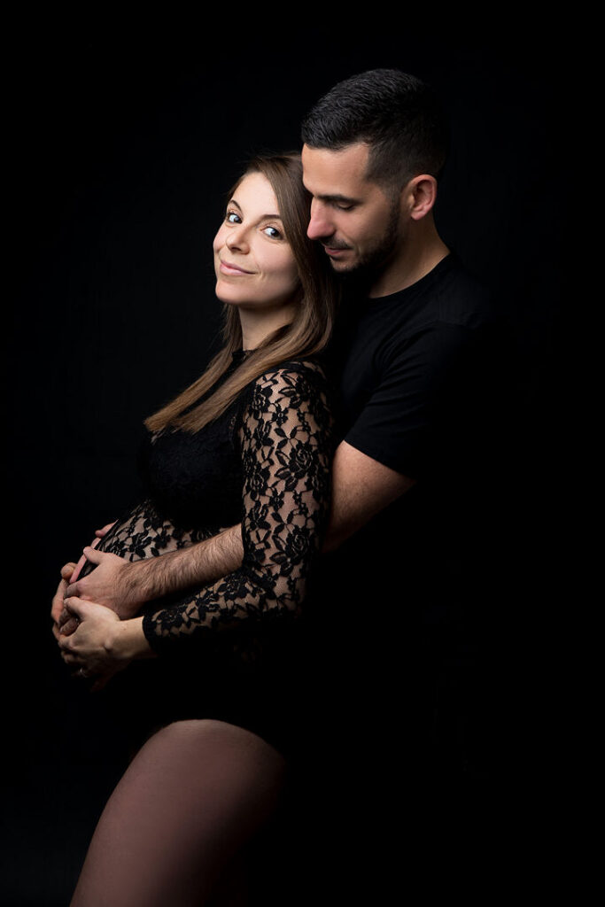 Une femme enceinte en robe de dentelle noire, berçant son ventre rond dans les mains de son partenaire. Une séance photo de couple sur fond sombre, immortalisant un moment de tendresse.