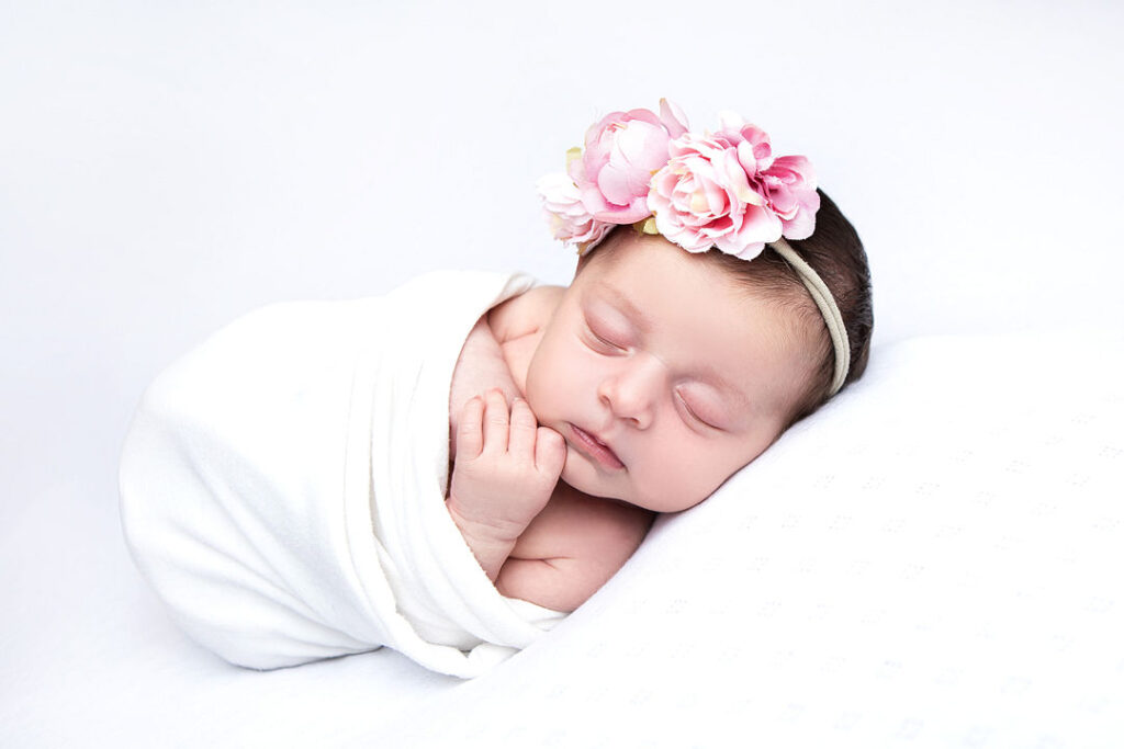 Un nouveau-né endormi, enveloppé dans une couverture blanche et coiffé d'une couronne de fleurs roses, est parfait pour une séance photo de nouveau-né. Photos paisibles d'un bébé lors d'une séance photo de nouveau-né.